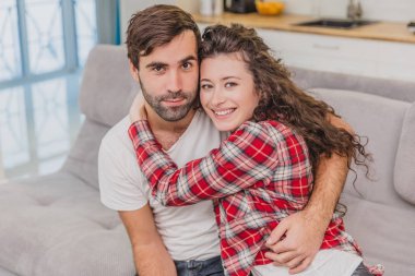 Genç çift kendi dairesinde rahatlatıcı, kanepede oturan, TV izlerken. aşıklar ve mutlu sarılmak. Doğrudan kameraya bak ve içtenlikle gülümse.