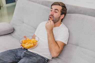 Tişörtlü adam kanepede oturuyor, cips yiyor ve televizyon izliyor. Kumandayı elinde tutarken. Evde kişilik.