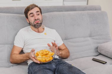 Tişörtlü adam kanepede oturuyor, cips yiyor ve televizyon izliyor. Kumandayı elinde tutarken. Evde kişilik.