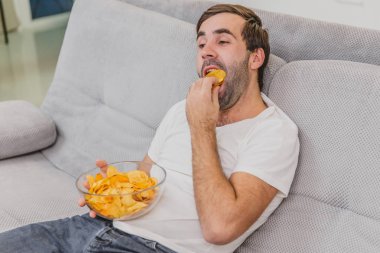 Tişörtlü adam kanepede oturuyor, cips yiyor ve televizyon izliyor. Kumandayı elinde tutarken. Evde kişilik.
