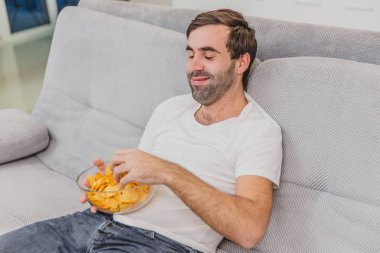 Tişörtlü adam kanepede oturuyor, cips yiyor ve televizyon izliyor. Kumandayı elinde tutarken. Evde kişilik.