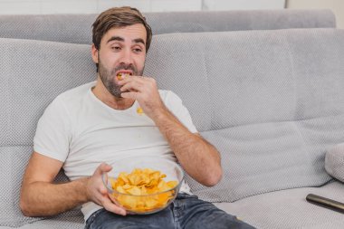 Tişörtlü adam kanepede oturuyor, cips yiyor ve televizyon izliyor. Kumandayı elinde tutarken. Evde kişilik.