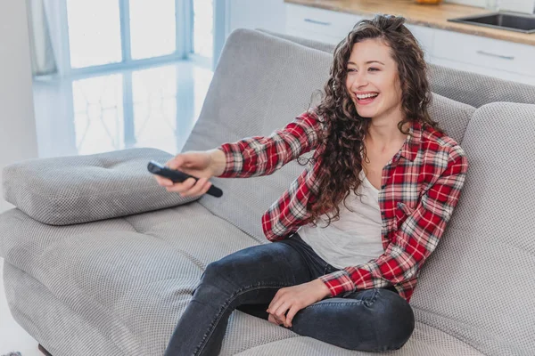 A beautiful woman watches the TV and is sitting on the couch and holds the remote control in his hand. A brunette in a shirt looks at the TV with long hair.