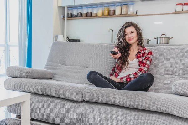 Eine schöne Frau schaut den Fernseher und sitzt auf der Couch und hält die Fernbedienung in der Hand. eine Brünette im Hemd schaut mit langen Haaren auf den Fernseher. — Stockfoto