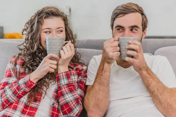 Una hermosa pareja de jóvenes. La gente graciosa levantó la taza de café con la cara cerrada. Durante esto, miran a la cámara y sonríen. Sentado en el sofá en casa . —  Fotos de Stock