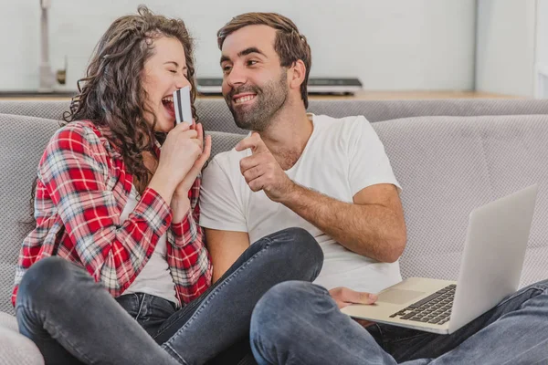 Casal bonito está fazendo compras on-line usando um laptop e sorrindo enquanto sentado no sofá em casa, mulher está segurando um cartão de crédito . — Fotografia de Stock