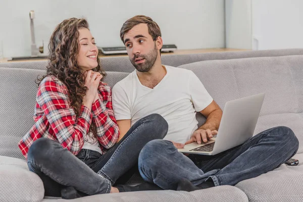 Гарна пара під час покупки в Інтернеті, використовуючи ноутбук і посміхаючись, сидячи на дивані вдома. Жінка просить чоловіка зробити покупку в Інтернеті . — стокове фото
