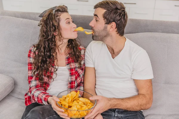 Leende hustru och man sitter på soffan. Titta på TV medan du äter chips i sängen. — Stockfoto