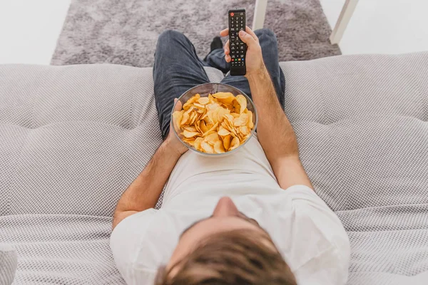 The guy in the T-shirt sits on the couch, eats chips and watches the TV. While holding the remote in his hands.