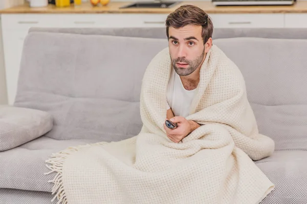 A man with a cold sitting on the couch in a blanket is as cold as possible. Man at home - sick man. Gray sofa in the room. The concept of a misfortune. The concept of health and healthy lifestyle.
