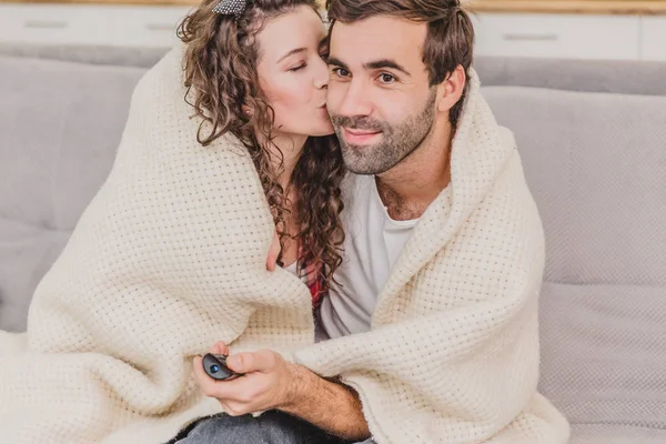 Молодая пара отдыхает в своей квартире, сидит на диване, смотрит телевизор. Покрытые одеялом, счастливые объятия . — стоковое фото