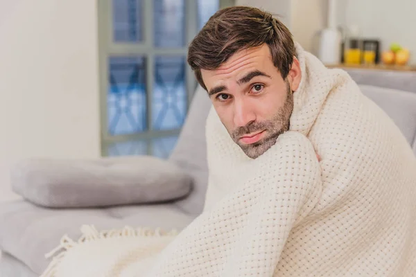 一个感冒的人坐在沙发上，裹着毯子，是尽可能冷的。男人在家 - 病人。房间里的灰色坐。不幸的概念。健康与健康的生活方式理念. — 图库照片