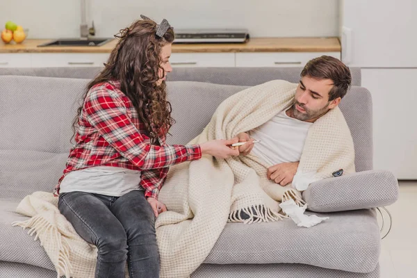 A man with a cold sitting on a sofa in a blanket blanket. Checking the temperature. The wife is sitting next to her and helps. Man at home. Sick young man Gray sofa in the room. Unhappy guy. Concept