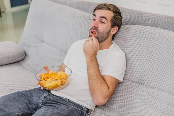 Killen i T-shirt sitter på soffan, äter chips och tittar på TV. Medan du håller fjärrkontrollen i händerna. Personlighet hemma. — Stockfoto