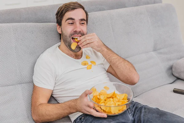 Tişörtlü adam kanepede oturuyor, cips yiyor ve televizyon izliyor. Kumandayı elinde tutarken. Evde kişilik.
