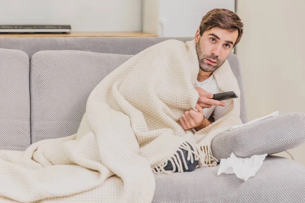 一个感冒的人坐在一个浅毯子的靠着的毯子的架子上。在家的男人是个年轻人。房间里的灰色坐。不幸的疾病概念. — 图库照片