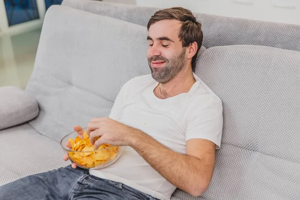 Killen i T-shirt sitter på soffan, äter chips och tittar på TV. Medan du håller fjärrkontrollen i händerna. Personlighet hemma. — Stockfoto