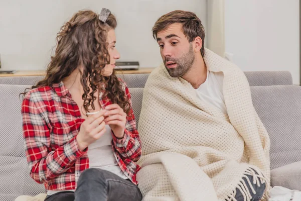 A man with a cold sitting on a sofa in a blanket blanket. Checking the temperature. The wife is sitting next to her and helps. Man at home. Sick young man Gray sofa in the room. Unhappy guy. Concept