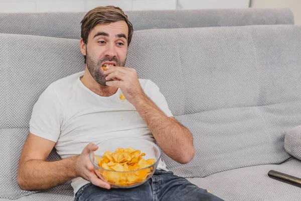 Tişörtlü adam kanepede oturuyor, cips yiyor ve televizyon izliyor. Kumandayı elinde tutarken. Evde kişilik.