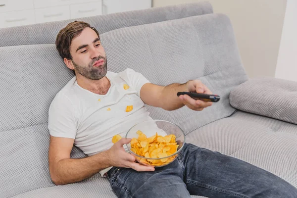 The guy in the T-shirt sits on the couch, eats chips and watches the TV. While holding the remote in his hands. Personality at home.