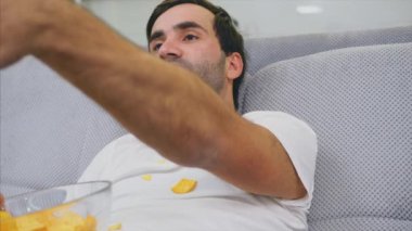 Tişörtlü adam kanepede oturuyor, cips yiyor ve televizyon izliyor. Kumandayı elinde tutarken. Evde kişilik. Yeşil ekran.