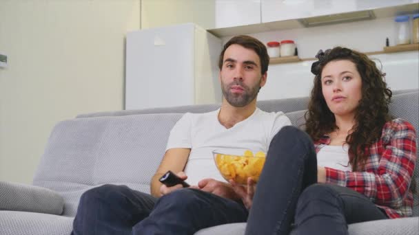 Lachende vrouw en man zittend op de Bank. Kijk naar de tv terwijl je chips eet in bed. Gekleed in zwarte denim broek. — Stockvideo