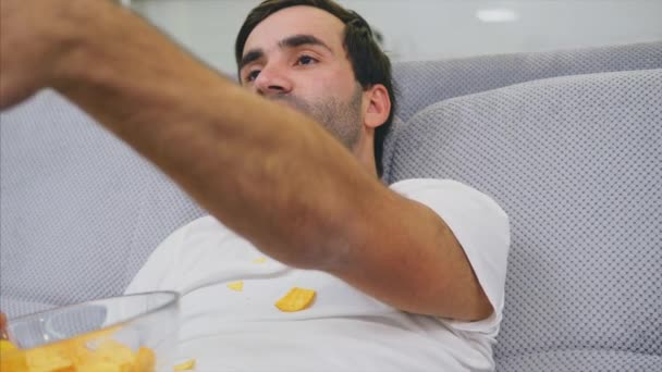 Killen i T-shirt sitter på soffan, äter chips och tittar på TV. Medan du håller fjärrkontrollen i händerna. Personlighet hemma. Grön skärm. — Stockvideo