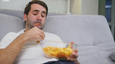 Tişörtlü adam kanepede oturuyor, cips yiyor ve televizyon izliyor. Kumandayı elinde tutarken. Evde kişilik.