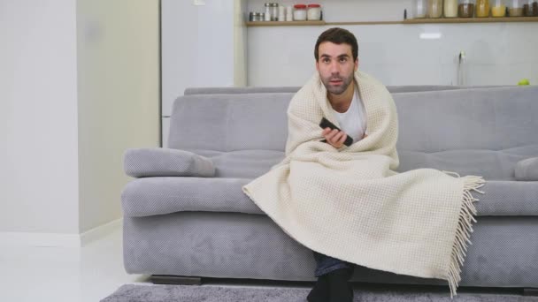 Un hombre con un resfriado sentado en el sofá en una manta es lo más frío posible. Hombre en casa - hombre enfermo. Sofá gris en la habitación. El concepto de una desgracia. El concepto de salud y estilo de vida saludable . — Vídeo de stock