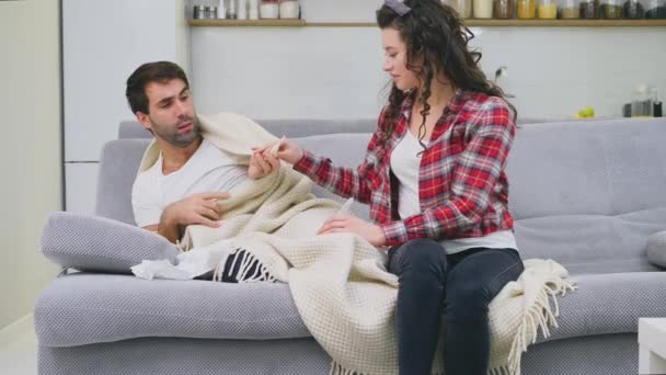 Ein erkälteter Mann sitzt in einer Decke auf einem Sofa. Kontrolle der Temperatur. die Frau sitzt neben ihr und hilft. Mann zu Hause. Kranker junger Mann graues Sofa im Zimmer. unglücklicher Typ. Konzept — Stockvideo