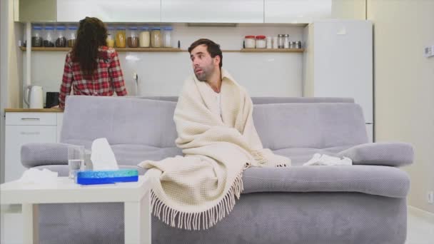 Un homme avec un froid assis sur un canapé dans une couverture. Je vérifie la température. La femme est assise à côté d'elle et aide. Un homme à la maison. Jeune homme malade Canapé gris dans la chambre. Un gars malheureux. Concept — Video