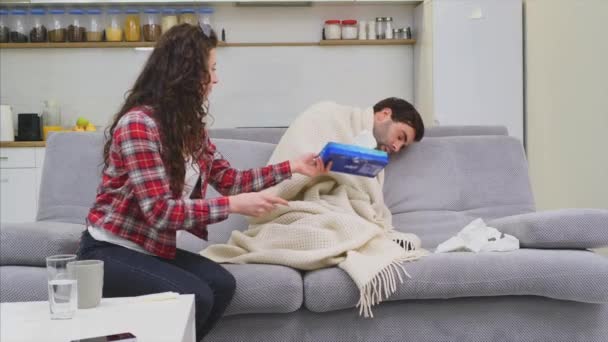 Um homem com um resfriado sentado em um sofá em um cobertor. A verificar a temperatura. A esposa está sentada ao lado dela e ajuda. Um homem em casa. Jovem doente Sofá cinzento no quarto. Um tipo infeliz. Conceito — Vídeo de Stock