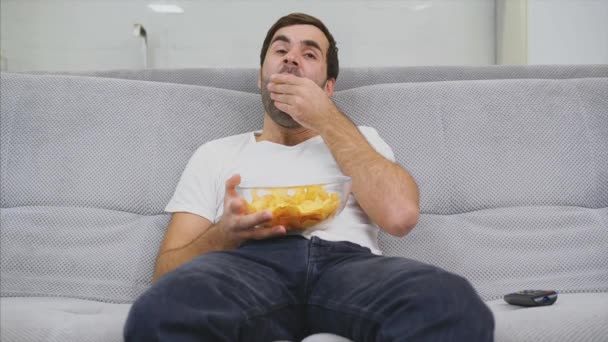 Killen i T-shirt sitter på soffan, äter chips och tittar på TV. Medan du håller fjärrkontrollen i händerna. — Stockvideo