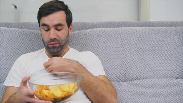 De man in het T-shirt zit op de Bank, eet chips en kijkt naar de TV. Terwijl hij de afstandsbediening in zijn handen houdt. — Stockvideo