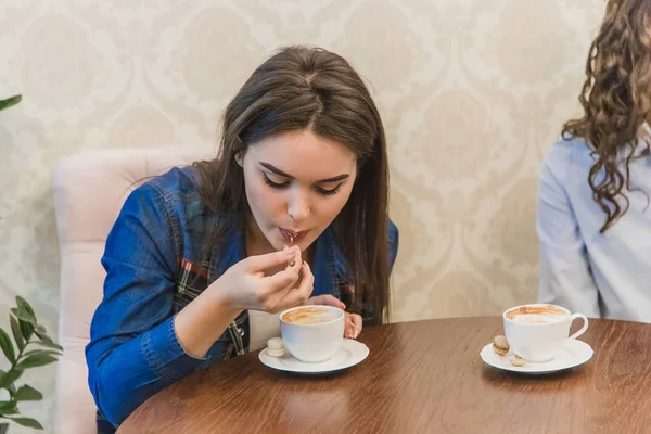 Mladá hezká dívka během setkání s přítelkyněmi, co sedí v kavárně. Pije cappuccino a přitom si ho užívá. — Stock fotografie