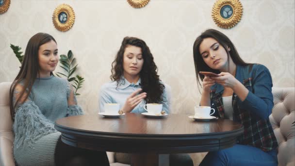 Tre amici seduti in un bar con uno smartphone e fare una conversazione divertente. Belle ragazze con lunghi bei capelli neri. Sul tavolo tre tazze di cappuccino . — Video Stock