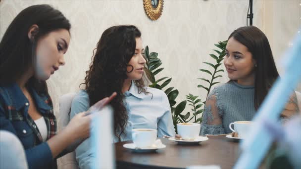 Tre venner sidder på en cafe med en smart telefon og har en sjov samtale. Gode piger med langt smukt sort hår. På bordet tre kopper cappuccino. Pigen opfanger begrebet – Stock-video