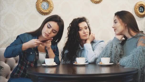 Tre amici seduti in un bar con uno smartphone e fare una conversazione divertente. Belle ragazze con lunghi bei capelli neri. Sul tavolo tre tazze di cappuccino . — Video Stock