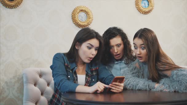 Três amigos, sentados em uma cafeteria usando um telefone inteligente e tendo uma conversa engraçada. Boas meninas com cabelo preto bonito longo . — Vídeo de Stock