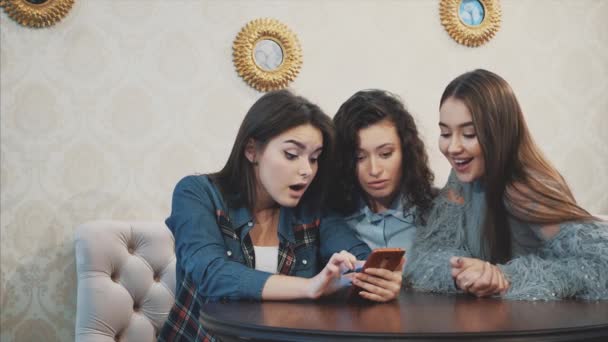 Tres amigos, sentados en una cafetería usando un teléfono inteligente y teniendo una conversación divertida. Buenas chicas con el pelo largo y hermoso negro . — Vídeos de Stock