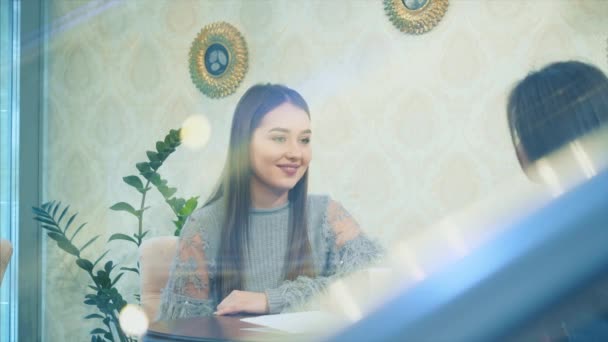 Menina bonita feliz senta-se em um café com suas namoradas. Conta uma história durante isto. Aproveitando o tempo juntos enquanto relaxa no café. Tem longo cabelo bonito preto . — Vídeo de Stock