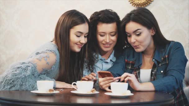 Trois amis assis dans un café avec un téléphone intelligent et avoir une conversation drôle. Bonnes filles avec de longs beaux cheveux noirs. Sur la table trois tasses de cappuccino . — Video