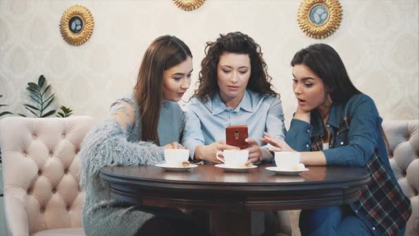 Drie vrienden zitten in een café met een slimme telefoon en een grappig gesprek. Goede meiden met lang mooi zwart haar. Op de tafel drie kopjes cappuccino. — Stockvideo