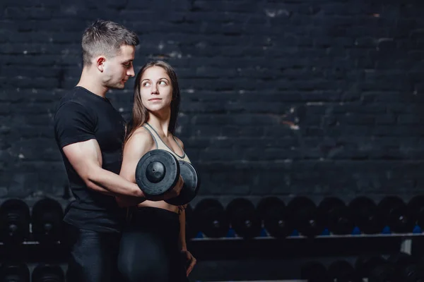 Two young guys and girls are engaged in the gym. During this time, they embracing themselves hold the weight in their hands while looking at each other.