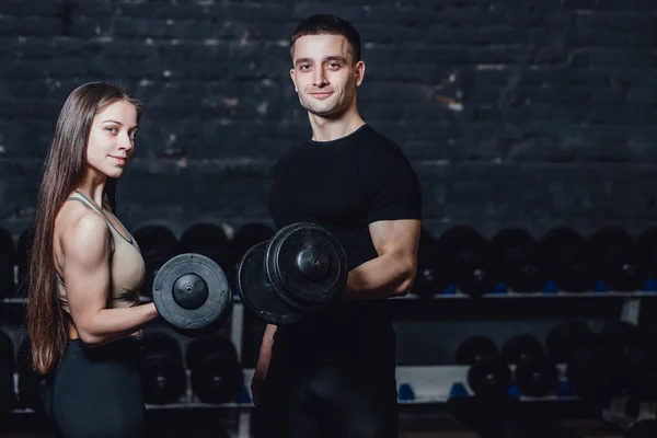 Two young guys and girls are engaged in the gym. During this they hold the weights in their hands while looking at the camera.