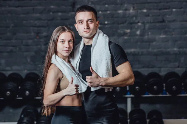 Jeune couple sportif homme et fille debout dans la salle de gym. Pendant ce temps, l'ordure blanche est maintenue sur le cou. Debout sur un fond noir et regardant la caméra . — Photo