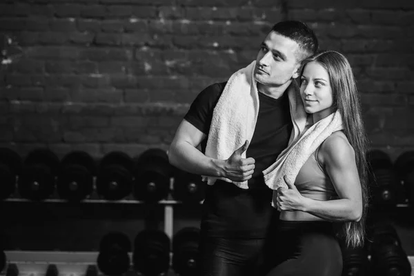 Jeune couple sportif homme et fille debout dans la salle de gym. Pendant ce temps, l'ordure blanche est maintenue sur le cou. Debout sur un fond noir et regardant loin . — Photo