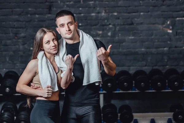 Jeune couple sportif homme et fille debout dans la salle de gym. Pendant ce temps, l'ordure blanche est maintenue sur le cou. Debout sur un fond noir et regardant loin . — Photo