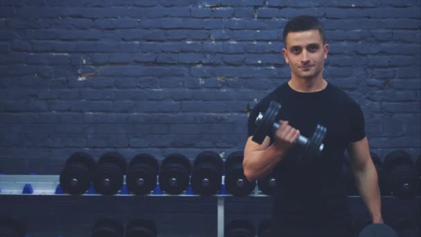 Sportig ung kille klädd i en svart T-shirt. En man med en idrotts kropp skakar musklerna i hantel. Under denna tid är han i gymmet. — Stockvideo