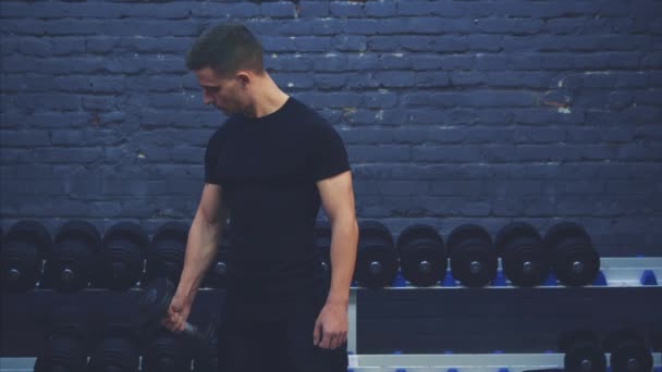 Un chico joven con una camiseta negra. Una gran barra en el gimnasio regularmente entrena las manos sobre un fondo oscuro . — Vídeo de stock